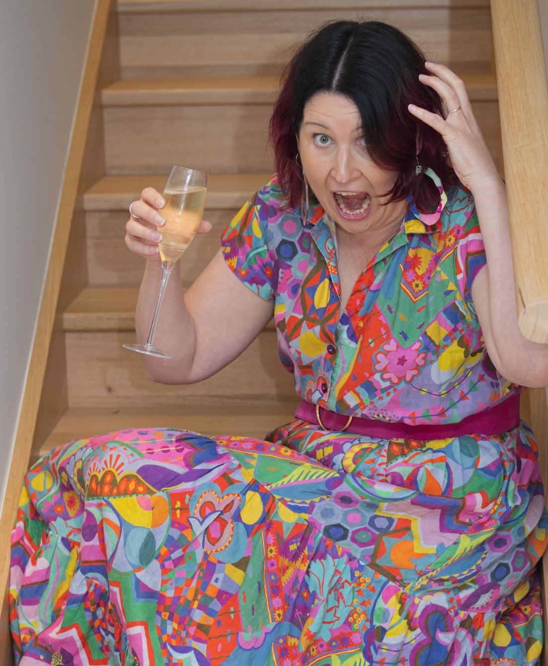 Woman in a brightly coloured dress sit on stairs drinking wine screaming in 'mock' frustration